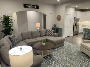 A modern living room with a sectional sofa, a coffee table, and a dining area with a kitchen in the background. - Dog friendly| community pool | gym| kayak| bayside (Selbyville)