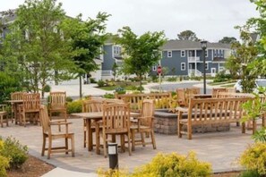 Un patio al aire libre con mesas y sillas de madera, rodeado de vegetación y edificios residenciales al fondo.