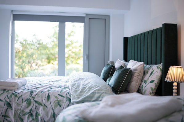 A bedroom with a bed, pillows, a bedside lamp, and a window with a view of greenery.