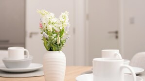 Una mesa de comedor con platos y tazas de cerámica blanca, un florero blanco con flores y una mesa de madera.