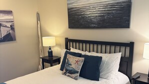 Un lit bien fait, recouvert d’une couverture bleue, un tableau représentant un paysage marin et une lampe sur une table de chevet.