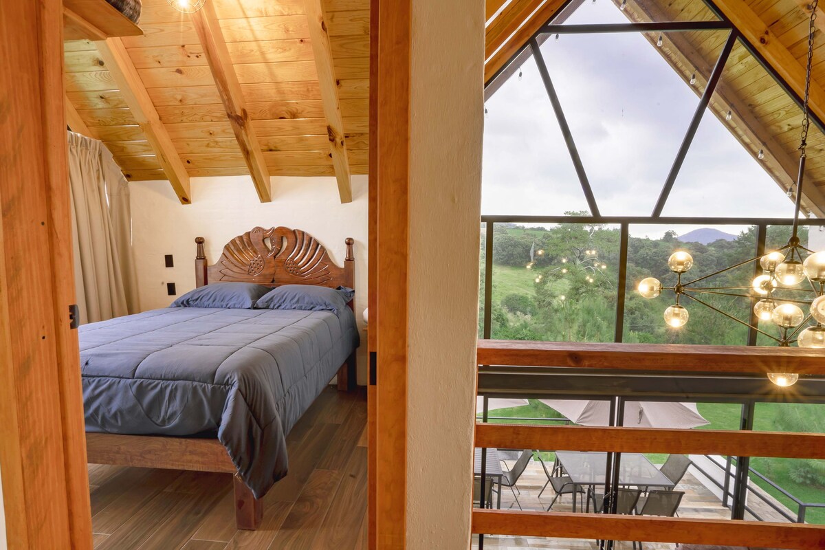 A bedroom with a wooden headboard, a bed with grey bedding, and a view of a green landscape through large windows.
