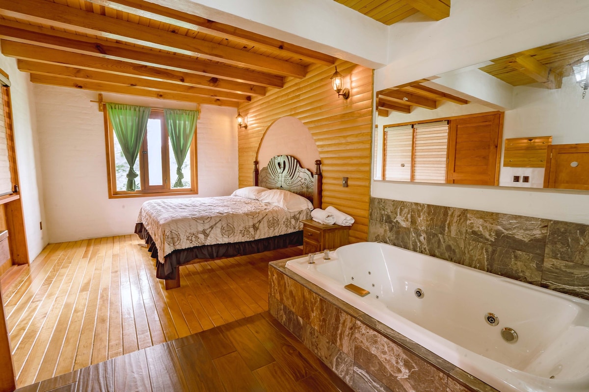 A bedroom with a large bed, a wooden ceiling, a window with curtains, a nightstand, and a bathroom with a bathtub and marble tiles.