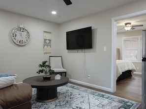 A living room with a round coffee table, a chair, a wall-mounted clock, a flat-screen TV, and a door leading to another room.