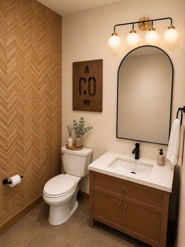 A bathroom with a herringbone wood floor, a white toilet, a wooden vanity with a mirror, and a wall-mounted light fixture.