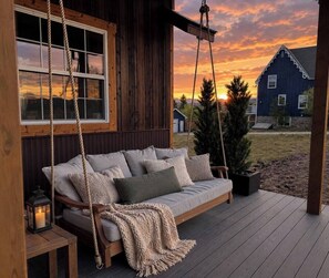 A wooden deck with a swing chair, a cushioned bench, and a small table with a lantern.