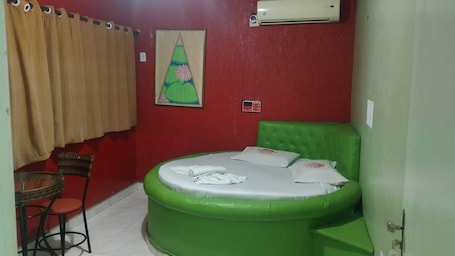 A small room with a green inflatable bed, a red wall, a window with beige curtains, a small table with a chair, and an air conditioning unit.. HOTEL TRÊS ESTRELAS