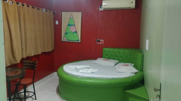 A small room with a green inflatable bed, a red wall, a window with beige curtains, a small table with a chair, and an air conditioning unit.