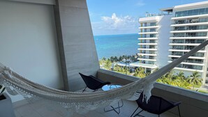 A balcony with a hammock, two chairs, and a view of the ocean and a hotel building. - Garza Blanca Resort: Spacious 1,830 sq ft 2-bedroom suite for up to 6 guests. (Cancún)
