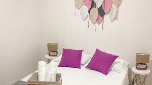 En snyggt bäddad säng med vita sängkläder och lila kuddar, ett sängbord med en lampa och en dekorativ detalj som hänger på väggen.