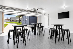 A garage with a large opening, a black TV mounted on the wall, and several black stools and tables arranged inside. - 1 block Lambeau, sleeps 14+, private yard, patio, heated tailgate garage, arcade (Green Bay)