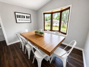 A dining room with a wooden table, white chairs, a potted plant, and a window with a view of houses. - 1 block Lambeau, sleeps 14+, private yard, patio, heated tailgate garage, arcade (Green Bay)