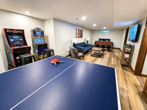 A ping pong table, two arcade machines, a sofa, a foosball table, and a TV in a room with wooden flooring and white walls. - 1 block Lambeau, sleeps 14+, private yard, patio, heated tailgate garage, arcade (Green Bay)
