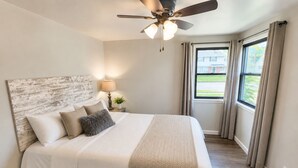 Un dormitorio con cama, ventilador de techo, ventana con cortinas y vistas al exterior.