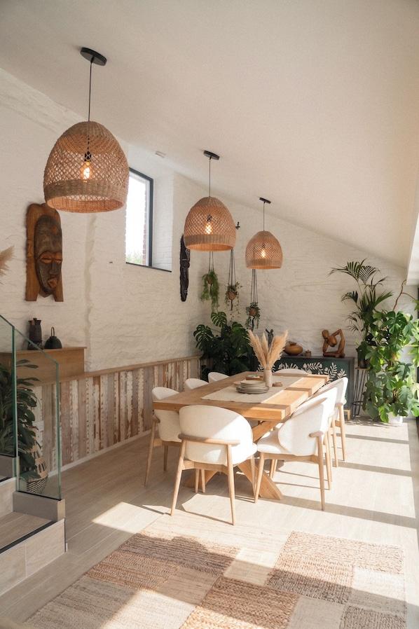 Un espace repas avec une table et des chaises en bois, des suspensions en osier et un escalier doté de rampes en verre.