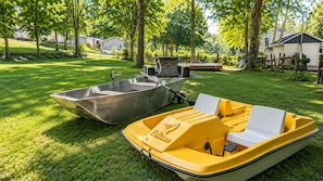 Ein gelbes und ein metallenes Boot auf einer grünen Wiese, im Hintergrund Bäume und Häuser.