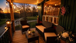 Une terrasse en bois, meublée de meubles en osier, décorée de guirlandes lumineuses et offrant une vue sur les arbres et une route.