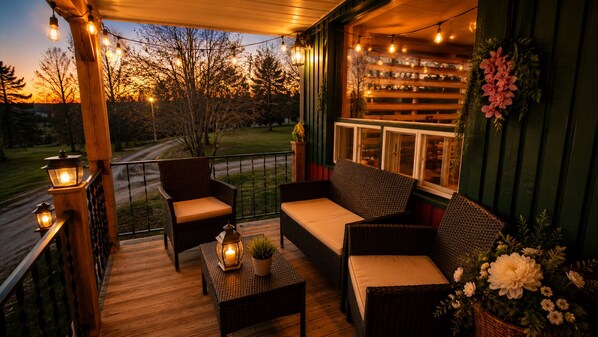A wooden deck with wicker furniture, string lights, and a view of trees and a road.