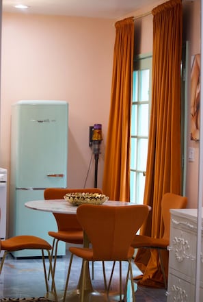 A dining area with a vintage teal refrigerator, orange chairs, a marble table, and a window with orange curtains.
