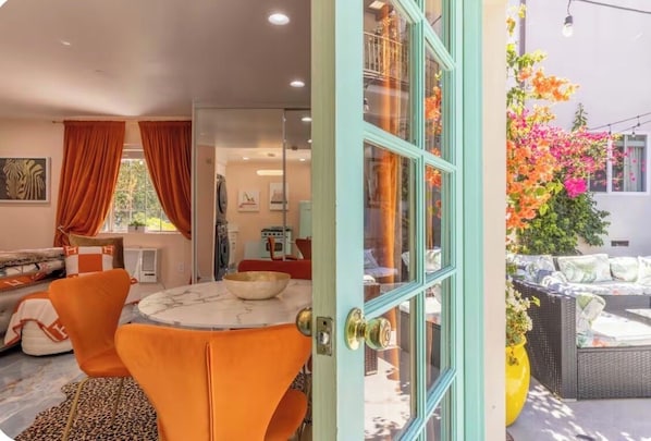 A dining area with a round table, orange chairs, and a view of the outdoor patio with wicker furniture and flowers.