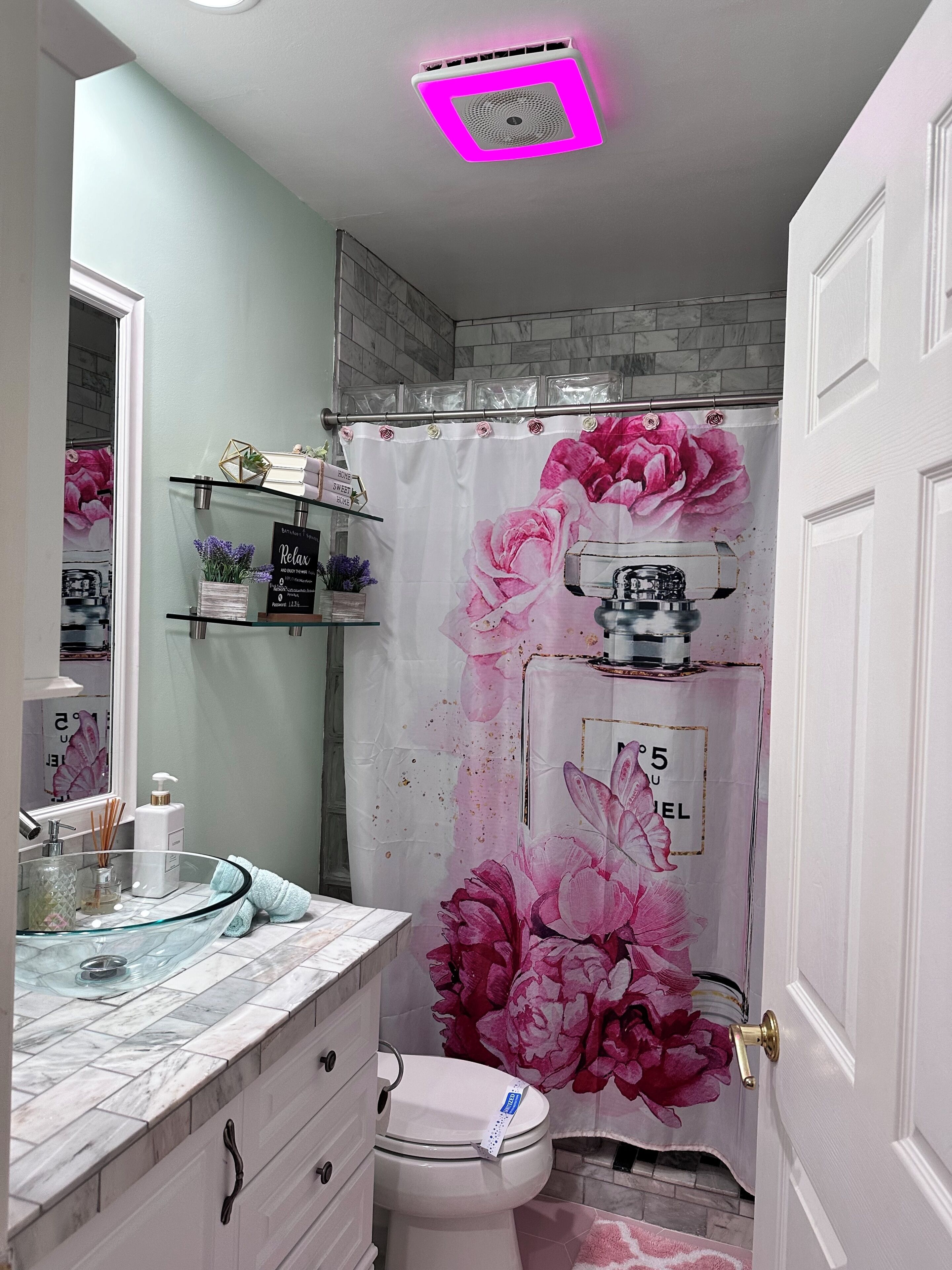 Une salle de bain avec un rideau de douche orné de motifs floraux, un lavabo en verre avec un plan de toilette en marbre et des toilettes.