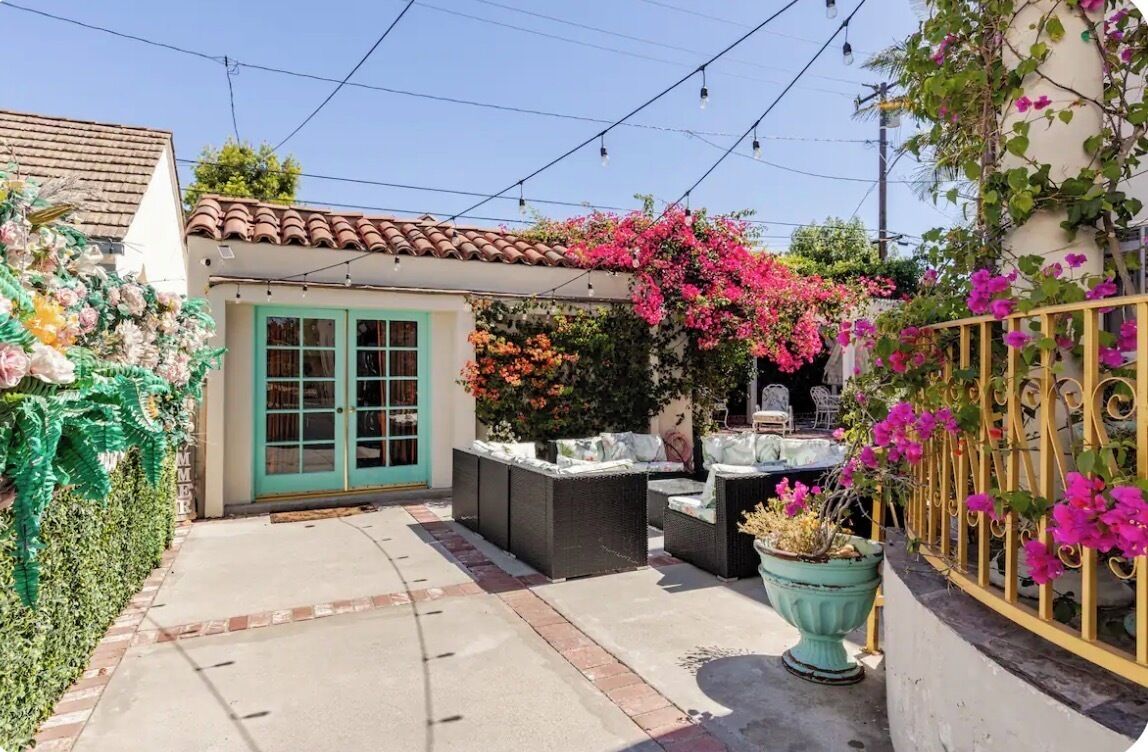 Une terrasse avec des meubles d’extérieur, un petit bâtiment avec une porte verte et des plantes en fleurs.
