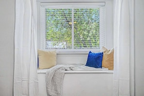 A window seat with a view of trees, white curtains, and two pillows.