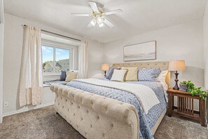 Quarto com cama grande, ventilador de teto, janela com cortinas e mesa de cabeceira com abajur e planta.