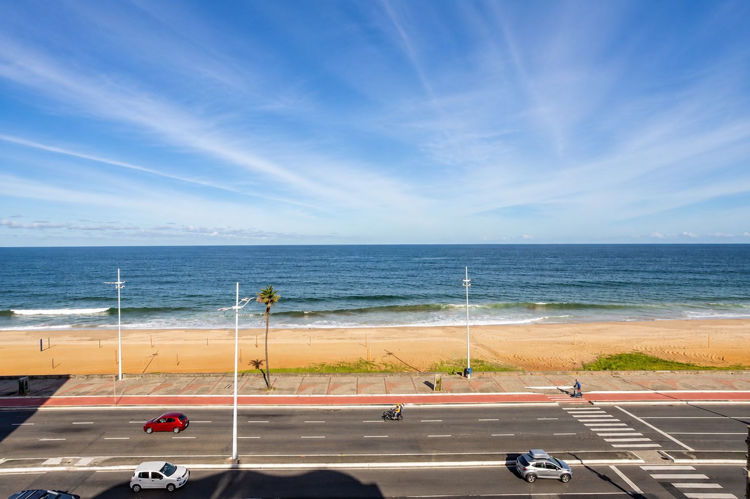 Vista de playa con una carretera en primer plano, cielo despejado y el océano al fondo.