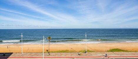 Vista de playa con una carretera en primer plano, cielo despejado y el océano al fondo.