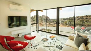 A modern living room with a flat-screen TV, red chairs, a glass coffee table, and a sofa with a view of a desert landscape through large windows. - Rock Cove By Homestead Modern (Pioneertown)