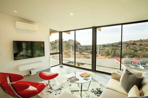 Un salón moderno con una televisión de pantalla plana, sillas rojas, una mesa de centro de vidrio y un sofá con vistas a un paisaje desértico a través de amplias ventanas.
