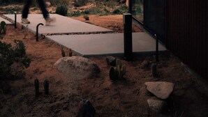 A desert landscape with a modern building featuring a glass entrance and a concrete pathway. - Rock Cove By Homestead Modern (Pioneertown)