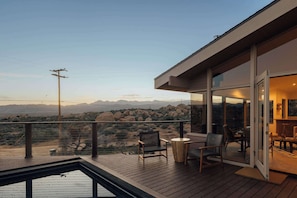 Eine Holzterrasse mit Sitzgelegenheiten im Freien, eine Glastür, die zu einem Innenraum führt, und Blick auf eine Wüstenlandschaft mit Bergen im Hintergrund.