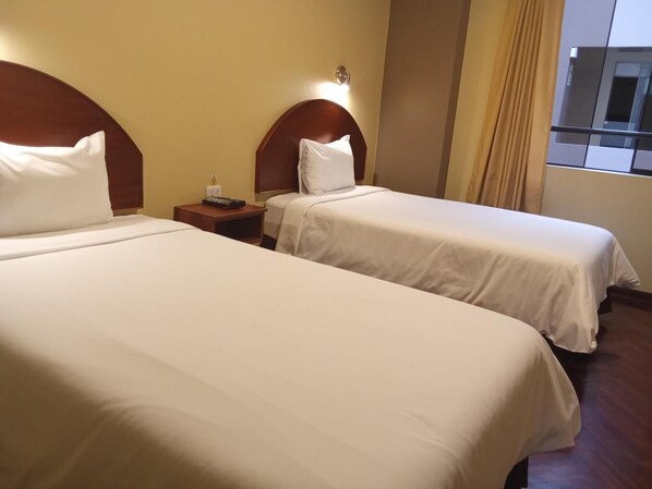 Una habitación de hotel con dos camas, una mesita de noche con lámpara y una ventana con cortinas.