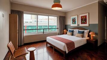A hotel room with a large bed, a wooden chair, a small table, and a view of the city through large windows.