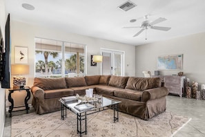 A living room with a brown leather sofa, a glass coffee table, a ceiling fan, and a view of palm trees through the windows. - Beachside Bliss with Paddleboards, Bikes & Island Fun - Mystic Mandalay - Roelens (Fort Myers Beach)