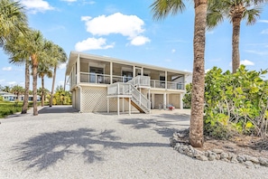 A two-story building with a balcony, surrounded by palm trees and a gravel driveway. - Beachside Bliss with Paddleboards, Bikes & Island Fun - Mystic Mandalay - Roelens (Fort Myers Beach)