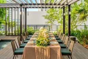 A long dining table set for a meal with chairs, a pergola overhead, and greenery in the background. - The Brickell Estates (Miami)