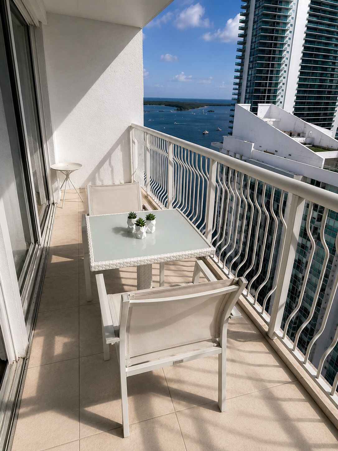 A balcony with a table and chairs, a small table with a plant, and a view of the sea and buildings.