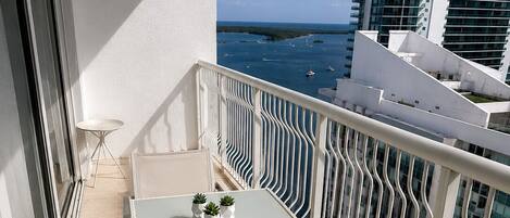 A balcony with a table and chairs, a small table with a plant, and a view of the sea and buildings.