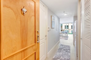 A hallway with a wooden door, white walls, a patterned rug, and a view of a building with windows. - The Brickell Estates (Miami)