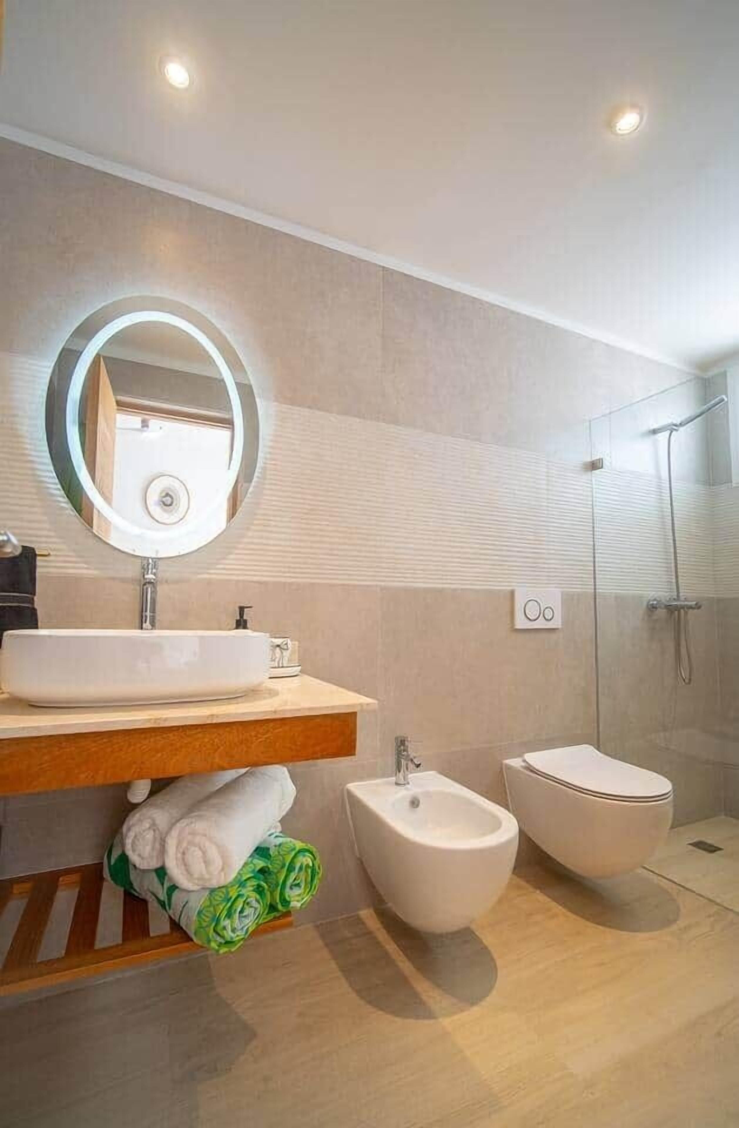A modern bathroom with a round mirror, a sink, a bidet, and a shower.