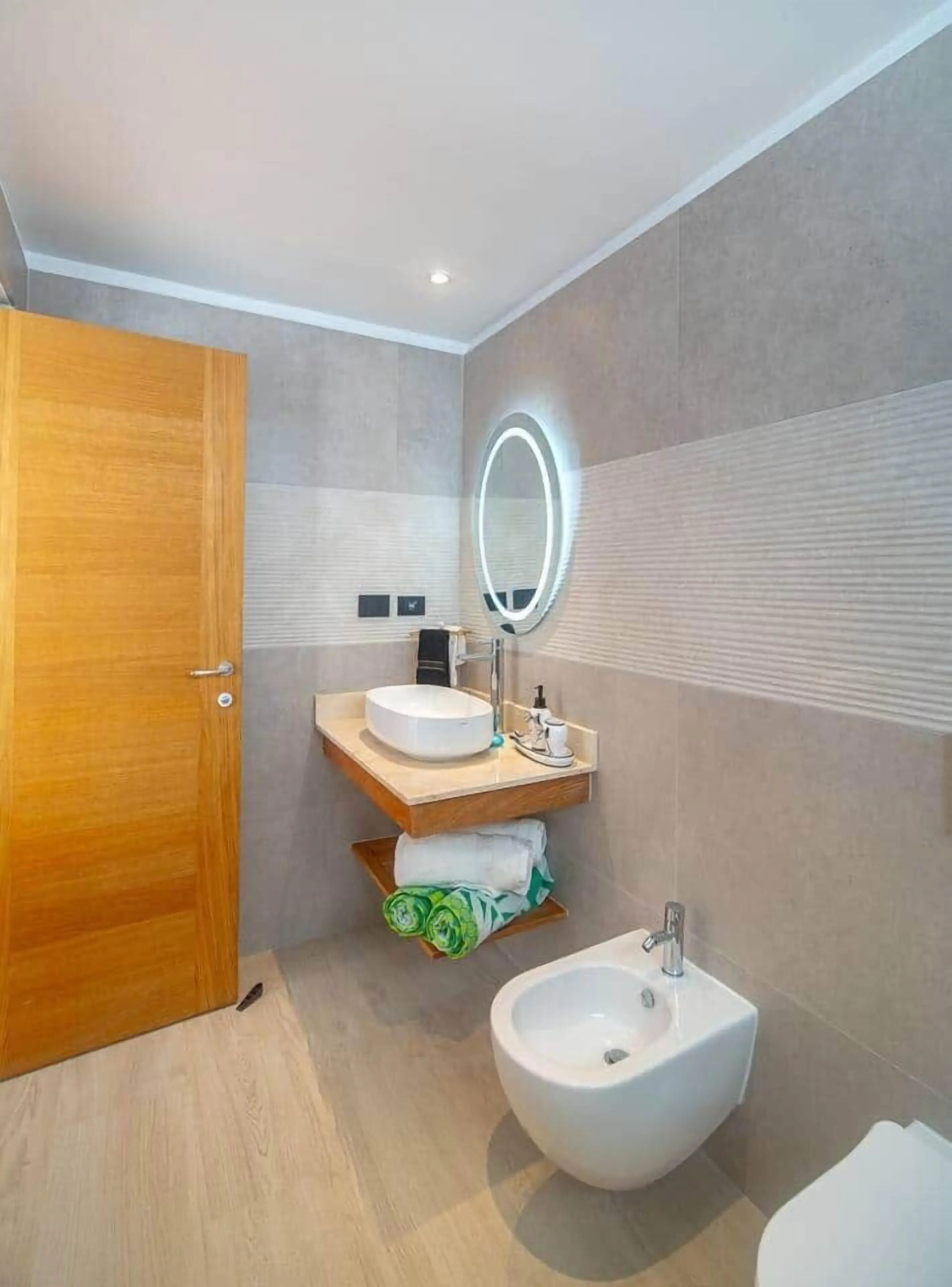 A modern bathroom with a wooden door, a vanity with a mirror, a bidet, and a towel rack.