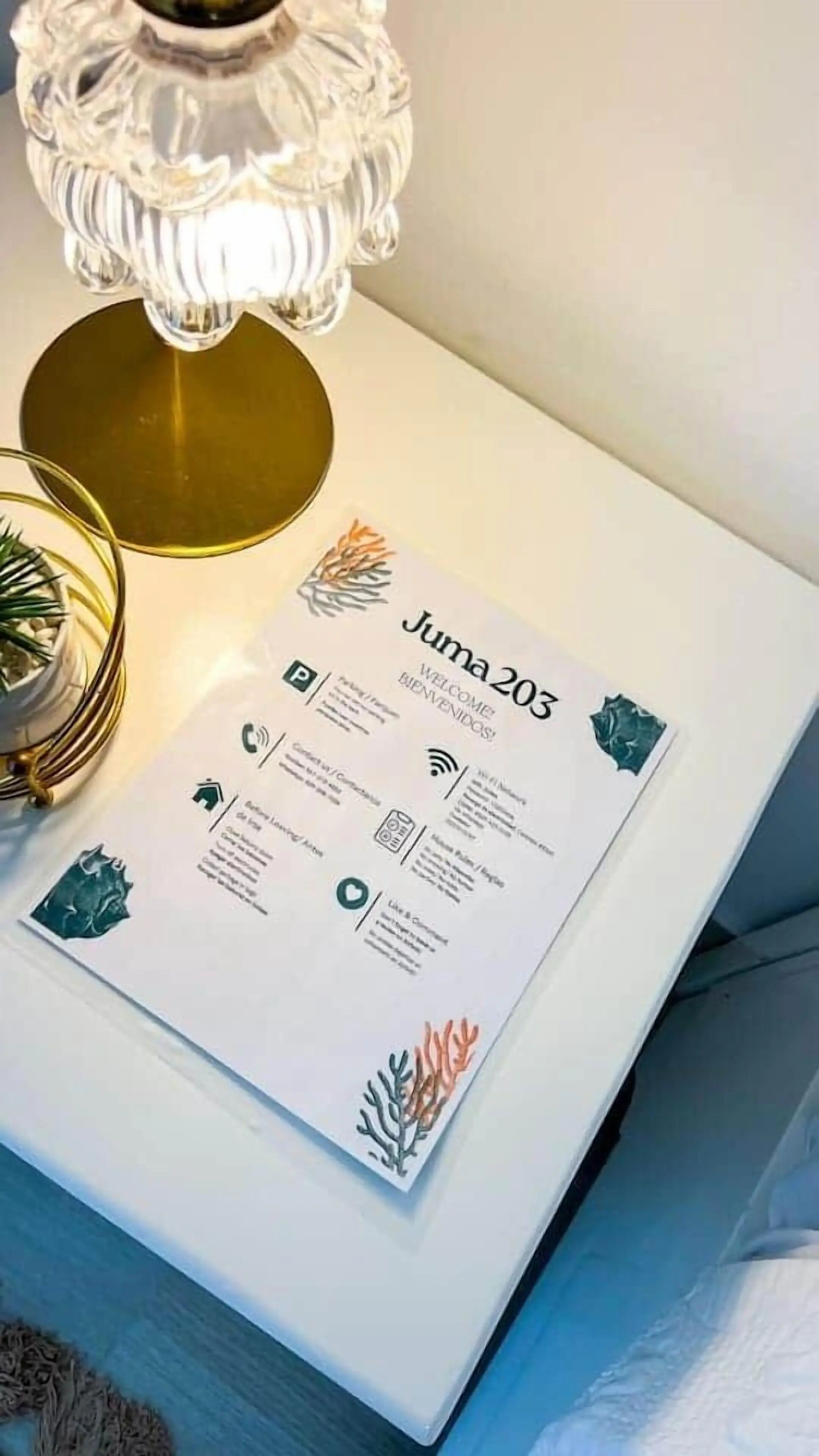 Une brochure de bienvenue intitulée « Juma 203 » est posée sur une surface blanche, à côté d’une lampe décorative et d’une petite plante.