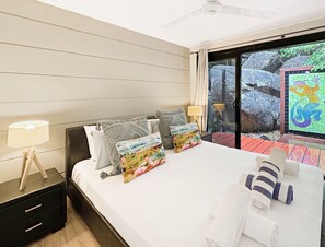 Habitació d'hotel amb llit, almoines, una tauleta de nit, una làmpada i vista a roques i vegetació a l'exterior.