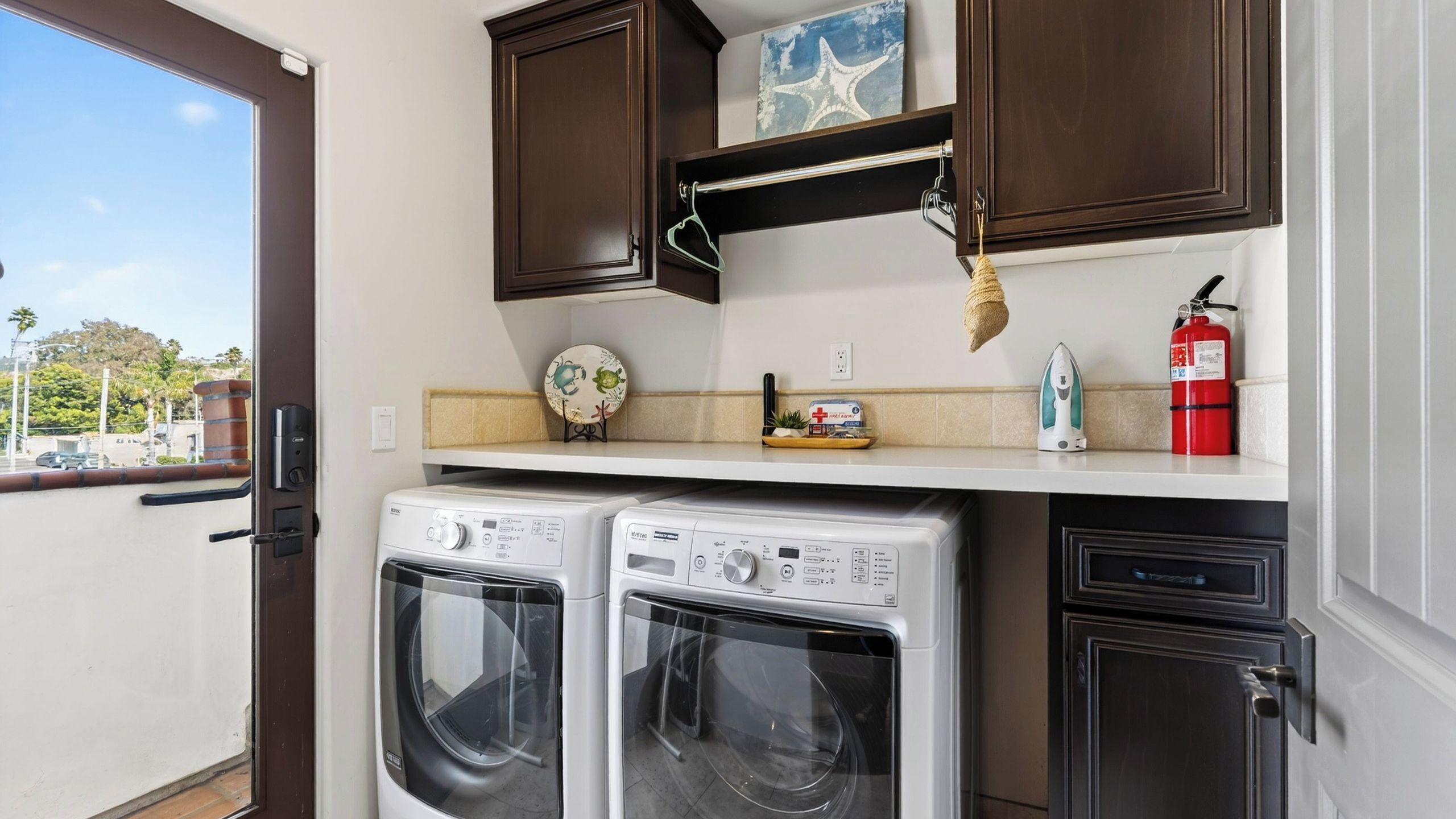 A laundry area with two front-loading washers, a shelf with a starfish decoration, a fire extinguisher, and a view of the outside.