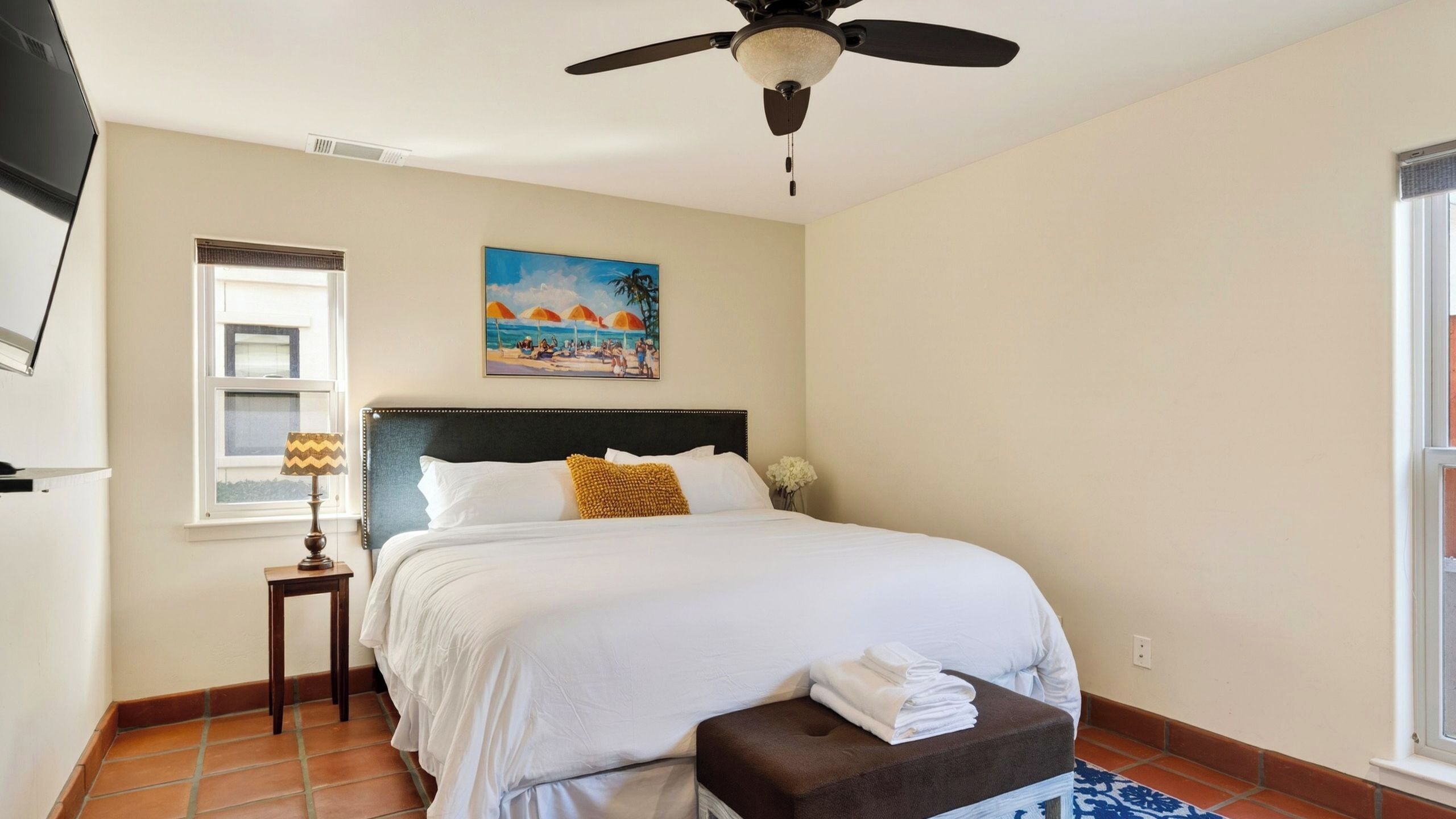 A bedroom with a bed, a ceiling fan, a window with blinds, a painting, and a nightstand.