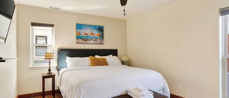 A bedroom with a bed, a ceiling fan, a window with blinds, a painting, and a nightstand.