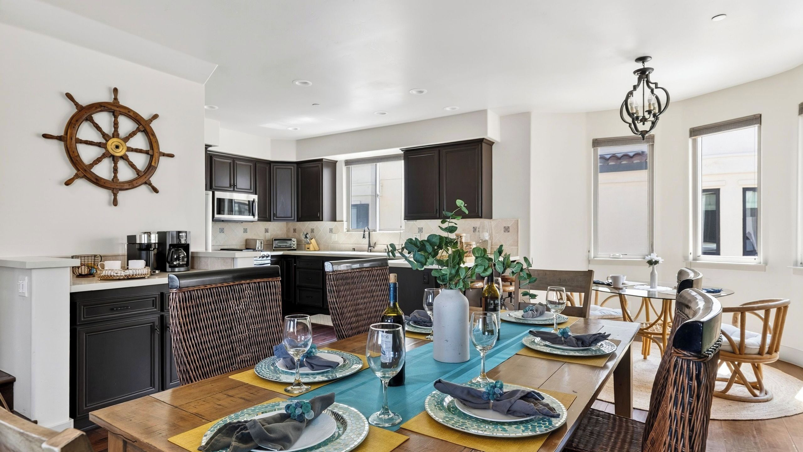 A modern kitchen with dark cabinets, a ship's wheel decoration, and a dining area set for a meal.